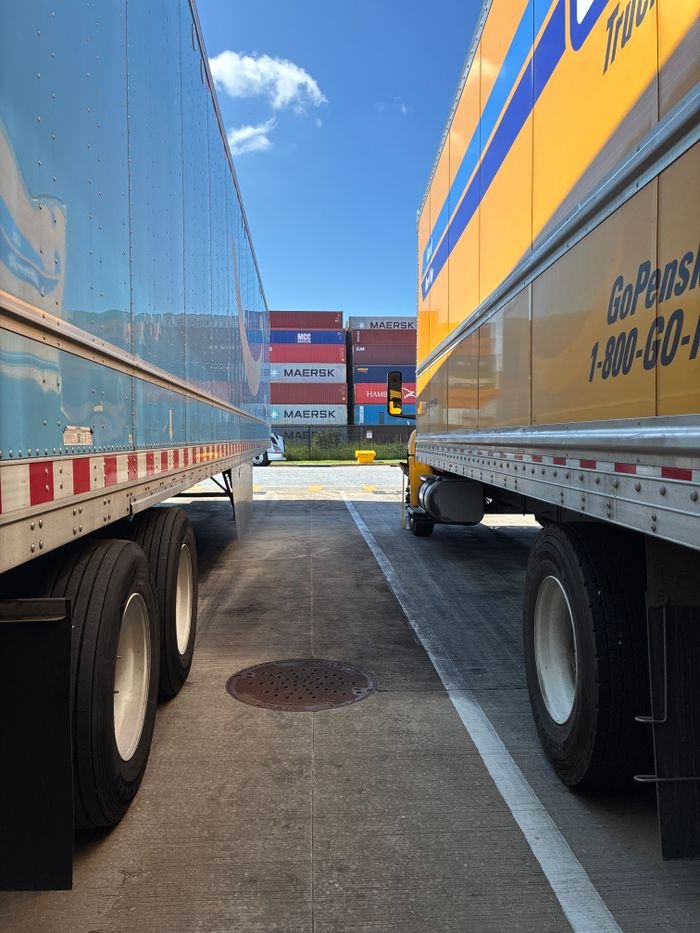 Wide shot of parked trailers with shipping containers in the background, clear sunny day.