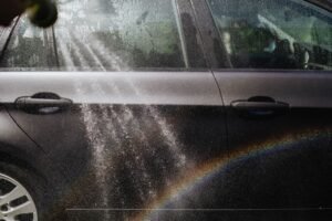 Close-up of a car being washed with a visible rainbow in sunlight.